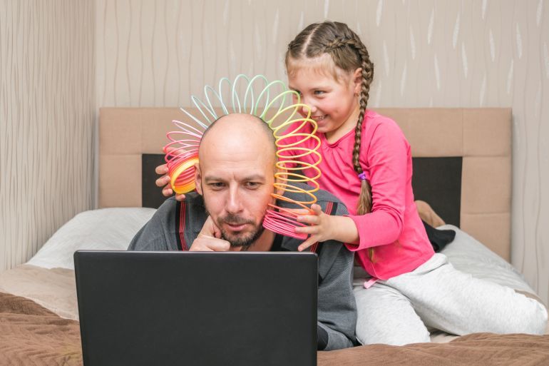 The daughter puts the slinky toy to the father's head. Family is quarantined at home. A little girl is making fun of dad.A child prevents a man from working on a laptop in his bedroom.