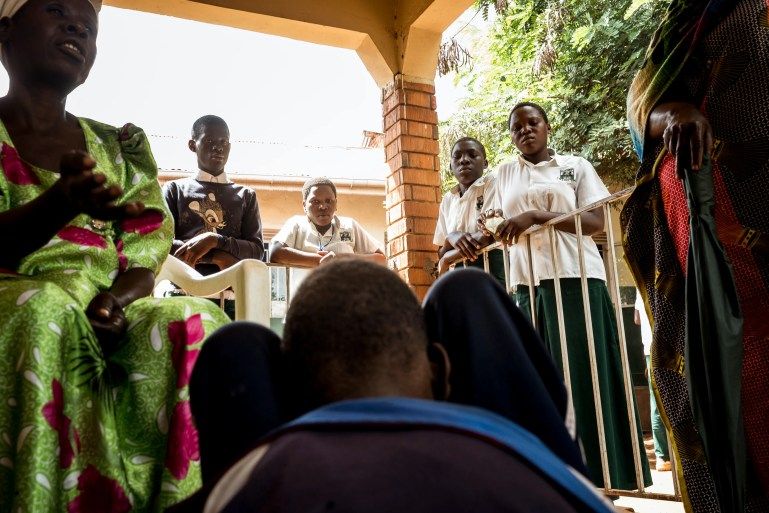 Students stare at Moses, 16, as his mother, Evelyn Mbwali, explains his story to a health worker at a local outreach clinic. Moses was brought to the clinic because his mother was worried about his lack of mobility [Christopher Hopkins/Al Jazeera]