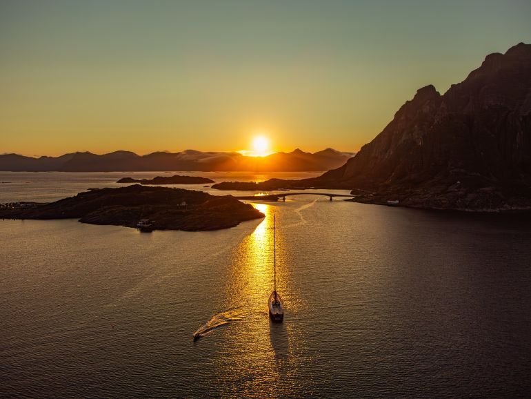 A sailboat under the midnight sun, behind a large hill in Lofoten, near Henningsvaer, in the distance tall mountains, sun setting.