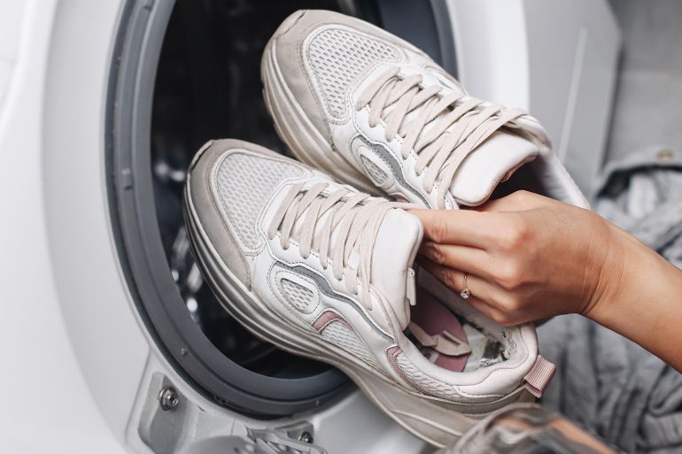 Woman putting white sneakers into washing machine. Home shoe cleaning routine. Laundry of dirty footwear. Washing fabric sports shoes. Freshness and hygiene. Modern household appliance use.