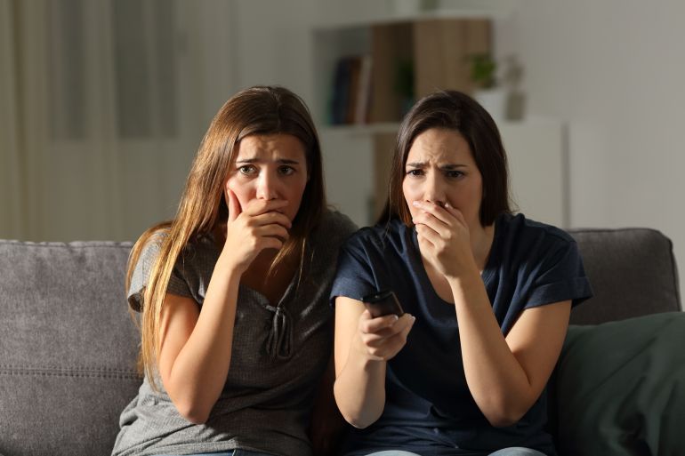 Front view portrait of two worried friends watching tv in the night sitting on a couch in the living room at home