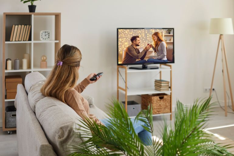 Back view of young woman relax on couch at home watch TV on lazy weekend. Female renter or tenant rest in living room enjoy film movie on television. Entertainment, relaxation concept.